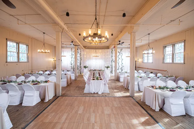 A beautifully decorated room with high white ceilings, white exposed brick walls and wood flooring. Multiple tables are lined up around the room draped in white table cloth with white fabric chairs. A chandelier hangs over the middle of the room and a dance floor is beneath it.
