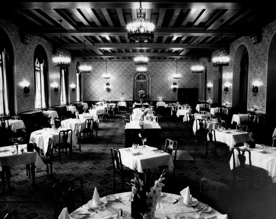 Une photo en noir et blanc d’une longue salle remplie de tables et de chaises. Les tables sont dressées de nappes blanches, de serviettes de table pliées et de multiples couverts d’argent. Des luminaires décoratifs sont suspendus au plafond.