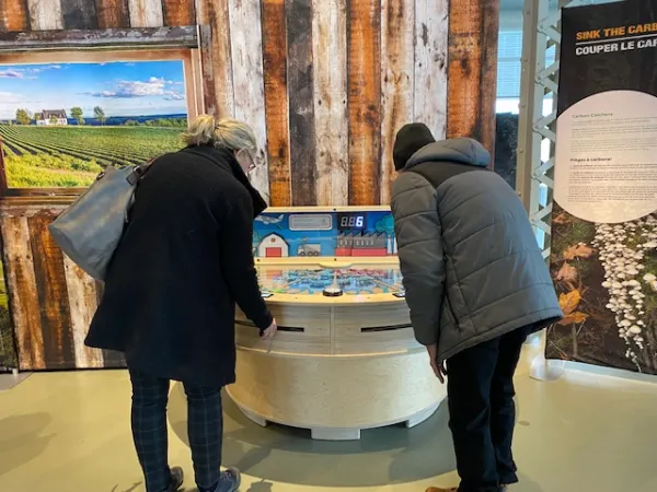 Two people standing facing what looks like a semi-circle wooden pin ball game. Each of the people are looking down at the top of the colourful table while pulling on levers just below the top of the table.