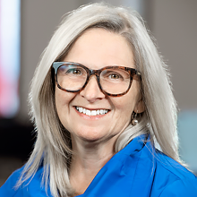 A portrait of Monique Horth, who has long grey hair, glasses and a blue blouse.