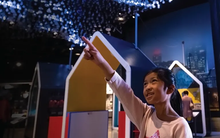 A girl is pointing at an exhibition as she stands next to a woman in front of an interactive display at the Canada Science and Technology Museum. Display cases are blurred out in the background.