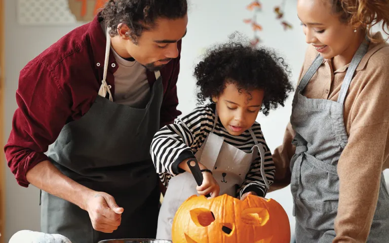 Two adults and a child stand over a pumpkin which has a face carved into it. The child is holding a large spoon and is scooping out the inside of the pumpkin while the adults watch. 