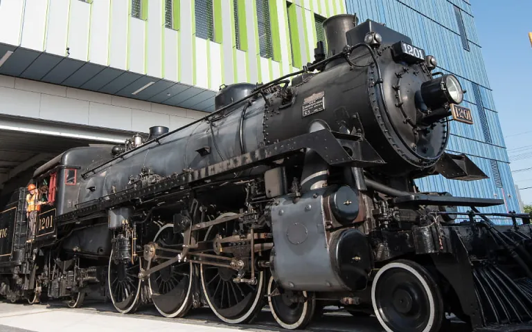 Steam locomotive CP 1201 is pushed by a diesel engine along tracks in a parking lot through a large door into the Ingenium Centre