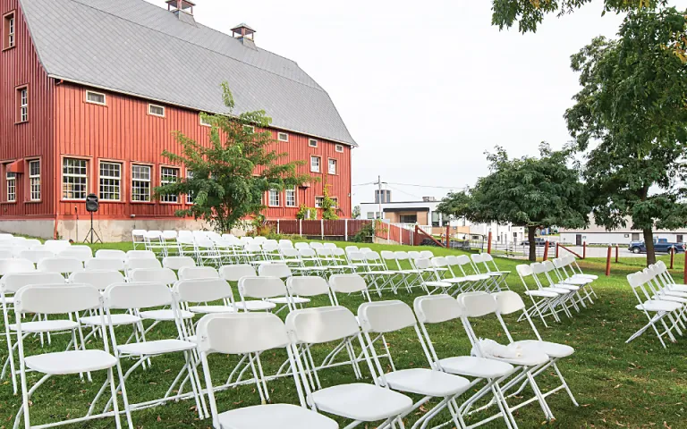 The outside of the barn at the Canadian Agriculture and Food Museum. The lawn is set up with rows of white fold-out chairs and has several trees surrounding the area. The entire barn is visible with the red walls, big windows, and grey roof giving a clean and minimal rustic look.