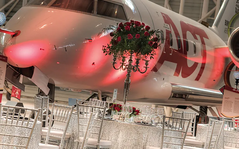 A view inside the Canada Aviation and Space Museum with a large white airplane in the background. Large tables are set up around the room with white translucent shimmering table cloths and elegant white chairs. Scattered around the room are classical Victorian-style lampposts with round lamp bulbs with geometric patterns on the glass.