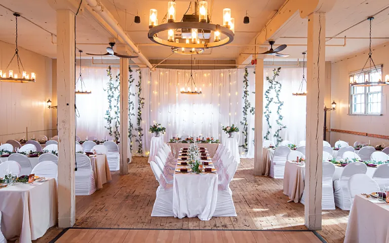 A beautifully decorated room with high white ceilings, white exposed brick walls and a wooden flooring. Multiple tables are lined up around the room draped in white table cloth with white fabric chairs. A chandelier hangs over the middle of the room where the space has been cleared out for a dance floor.