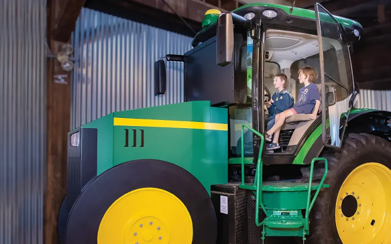 A wide shot of a climb-aboard tractor inside a museum space; two children are visible in the tractor cab.