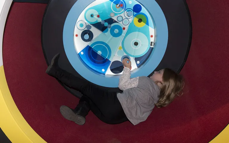 Child laying on her back inside an exhibit tube while spinning an interactive to see pieces move.
