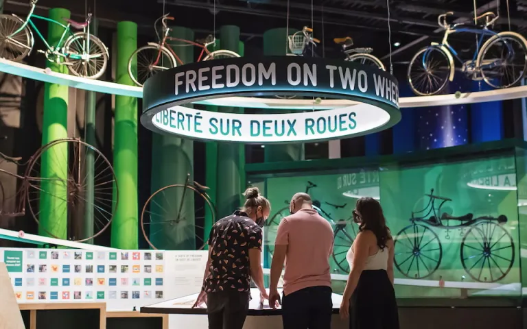 Un plan large montre trois personnes debout au milieu d'une exposition sur le thème du vélo. Diverses bicyclettes sont exposées sur de hautes étagères et à l'intérieur d'une vitrine en verre, et les mots "Liberté sur deux roues" sont visibles.