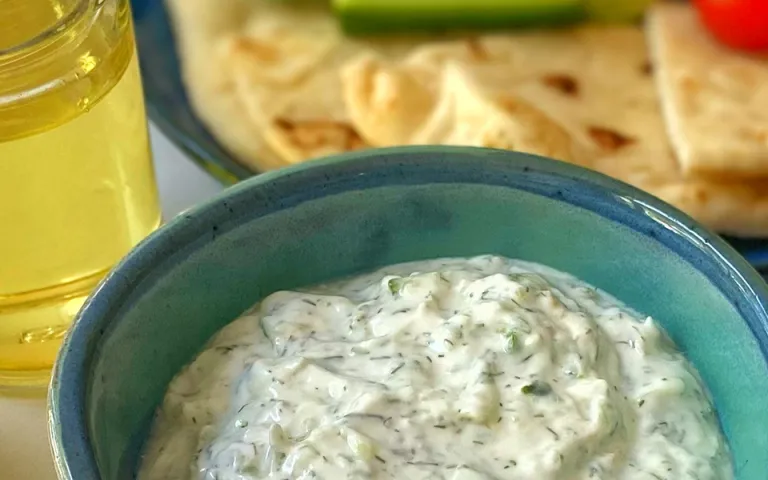 A blue bowl of dip sits on a white tabletop. Lemon wedges, olive oil and a salt bowl are visible around the dip. A blue plate with vegetables and pita wedges is visible in the background.  