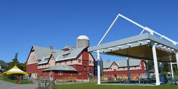 A wide shot of two large, red-and-white agricultural buildings and a navy blue silo. Green grass and a covered, outdoor picnic area are visible in the foreground, and a blue sky in the background.