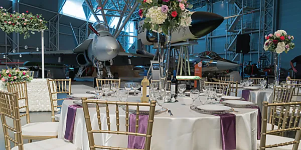 Grande salle avec des tables recouvertes de nappes blanches aux accents violets, et un avion blanc au fond. Chaque table est entourée d’élégantes chaises blanches et dorées. La pièce est décorée de fleurs aux couleurs pâles, vertes et roses.