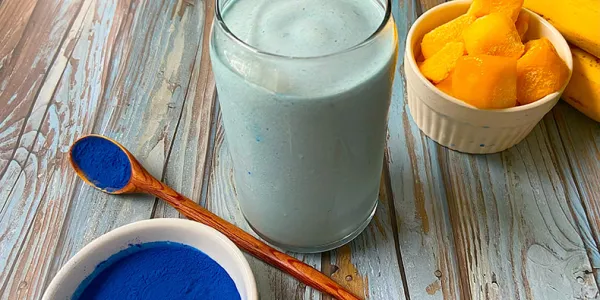 Un verre rempli d’une épaisse boisson bleu repose sur une surface de bois rustique. Un bol et une cuillère avec de la poudre bleue se trouvent à l'avant-plan, avec des mangues et des bananes à l'arrière-plan.