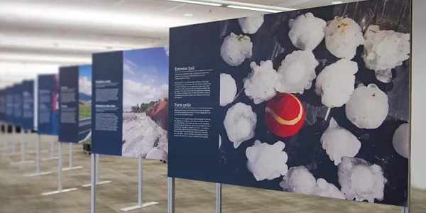 A wide shot of cascading exhibition photographic panels. The panel in the forefront is of a red tennis ball amongst many pieces of hail stones, the same size as the tennis ball.