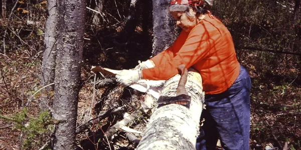Person in jeans wearing an orange shirt standing at a log. An axe lies on top of the log.