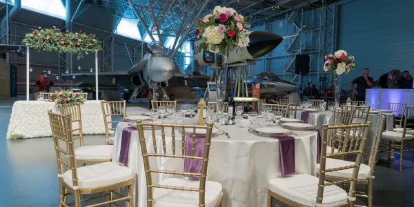 Grande salle avec des tables recouvertes de nappes blanches aux accents violets, et un avion blanc au fond. Chaque table est entourée d’élégantes chaises blanches et dorées. La pièce est décorée de fleurs aux couleurs pâles, vertes et roses.