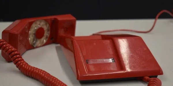 Un téléphone en plastique rouge posé sur une table gris clair, avec fond noir. Le combiné du téléphone, dans lequel se trouve le cadran rotatif, est posé sur la table à côté de la base de l’appareil, et est relié à celle-ci par un cordon rouge en spirale.