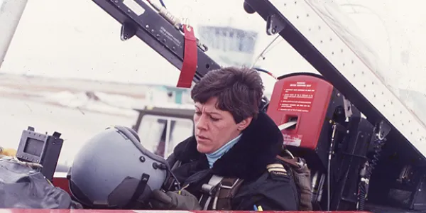 A woman sitting in the cockpit of a fighter jet with the canopy open, preparing to put on her flying helmet. 