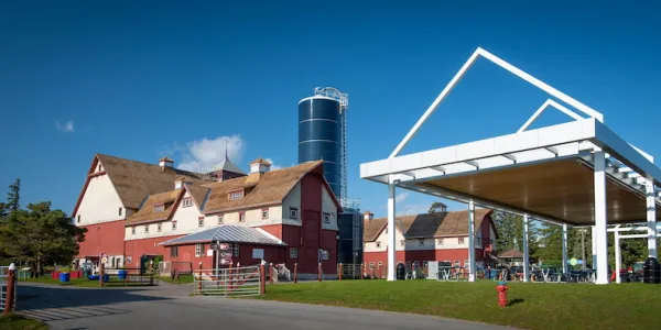 A wide shot of two large, red-and-white agricultural buildings and a navy blue silo. Green grass and a covered, outdoor picnic area are visible in the foreground, and a blue sky in the background.