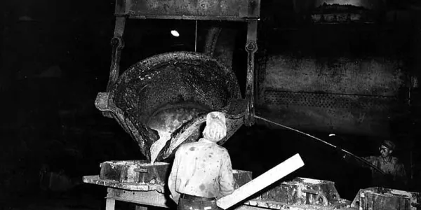 A black-and-white historical image shows a huge wooden machine with a large bucket. The bucket is tipping liquid metal into moulds below, and a man is overseeing the process with his back to the camera.