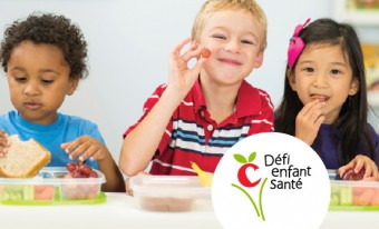 Trois jeunes enfants assis en file mangent leur dîner. Le garçon au centre sourit et tend un raisin. Le logo Défi enfant santé apparaît dans un cercle blanc au bas de la photo.
