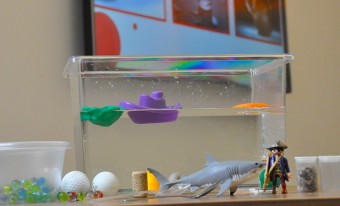 Un réservoir d'eau contient un bateau en plastique violet flottant et un rocher en plastique vert. À l'extérieur du réservoir se trouvent des objets épars tels qu'un requin en plastique, un pirate Playmobil et plusieurs balles de golf.