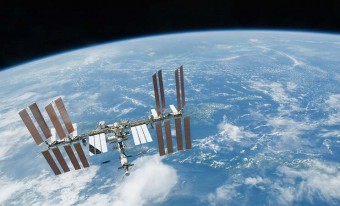 La Terre vue de la Station spatiale internationale. La lumière du soleil illumine les panneaux solaires de la SSI. La Terre est bleu pâle, et des nuages vaporeux en voilent la surface. On voit l’obscurité de l’espace en haut de l’image, au-dessus de la Terre.
