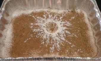 Un cratère formé de cacao et de farine, dans un plat rectangulaire jetable. Le cratère est brun et blanc, avec des traînées blanches laissées par la farine qui a jailli du centre. 