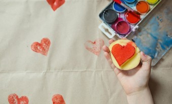 A child’s hand holds a potato stamp with painted red heart. In the background, a paint set and several stamped red hearts are visible against a white background
