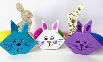 Trois têtes de lapin en origami (un bleu, un blanc et un mauve) sont alignées côte à côte sur un fond blanc. Chacun des lapins a un visage de style BD dessiné à l’encre noire. Derrière les lapins en papier, on aperçoit une collection d’œufs aux couleurs vives et des lapins en bois décoratifs.