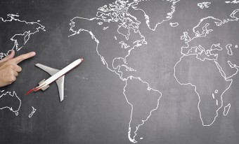 Modèle d'un avion de ligne, positionné sur un tableau noir sur lequel une carte du monde est dessinée à la craie blanche. À côté de l'avion se trouve une main avec un doigt qui indique la direction de vol de l'avion. 