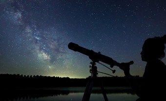 A silhouette of a woman with a telescope in the foreground is looking at the Milky Way galaxy in the night sky.
