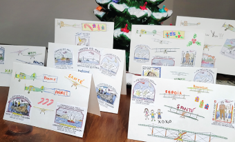 Huit cartes de vœux aux couleurs vives sont disposées sur une table de bois. Chaque carte présente des images différentes sur le thème de l'aviation. Les décors de Noël sont visibles à l’avant-plan et à l'arrière-plan de l'image.