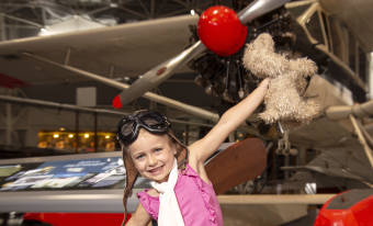 A smiling Caucasian child wearing a pink top, white scarf and a brown aviator hat and goggles on her head is holding a stuffed dog toy.  Her arms are extended as though she is flying.  Behind her is the front of a grey propeller plane with a red nose.