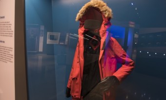 A red winter coat with brown fur trim on the hood inside of a glass display case.