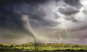 A tornado funnel is shown touching down onto green hilly farmland below a dark stormy sky with lightning bolts.  Various homes and barns can be seen in the distance.