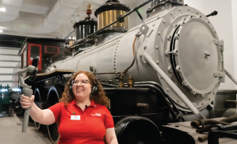  A museum employee stands in front of a large gray and black steam locomotive. She is wearing a headset and is smiling at a smartphone's screen held by a gimbal in her outstretched arm.