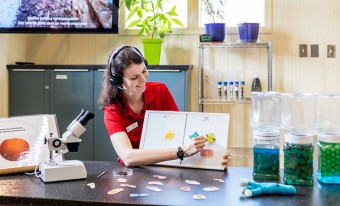 A museum staff member wearing a red shirt and headset sits at a lab counter, engaging in a hands-on science demonstration. She holds a whiteboard displaying colorful illustrations, explaining the concept of decomposition. A microscope, educational materials, and jars filled with green and blue beads are arranged on the counter, with plants and lab equipment visible in the background.