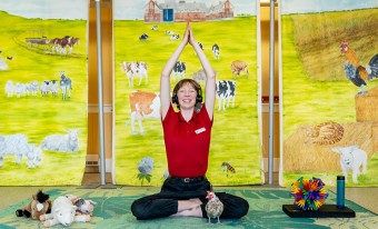 A museum staff member wearing a red shirt with black jeans and headset, is sitting cross-legged with her arms above her head and hands touching. She is sitting on a green mat with stuffed animals on the left and a colourful ball on the right. In the background there is a painted farm scene with animals and a barn.