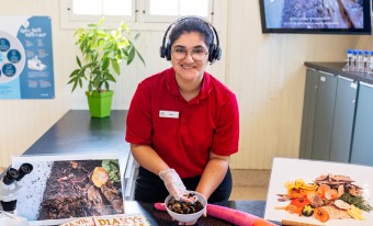 A museum staff member wearing a red shirt and headset sits at a lab counter holding a bowl with dirt and worms inside it. On the counter there is an image of earth with worms in it and another image with food for compost. In the background we see a plant and other lab equipment.