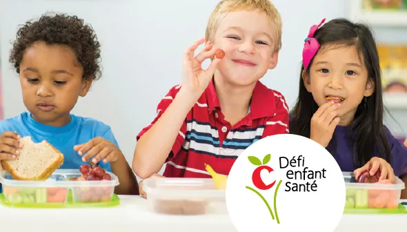 Trois jeunes enfants assis en file mangent leur dîner. Le garçon au centre sourit et tend un raisin. Le logo Défi enfant santé apparaît dans un cercle blanc au bas de la photo.