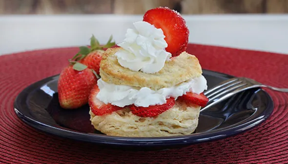 A fresh strawberry shortcake sits on a blue plate, on top of a placemat. The shortcake consists of a biscuit bottom, a strawberry and whipped cream middle, and another biscuit, with a whipped cream and juicy strawberry topping. 