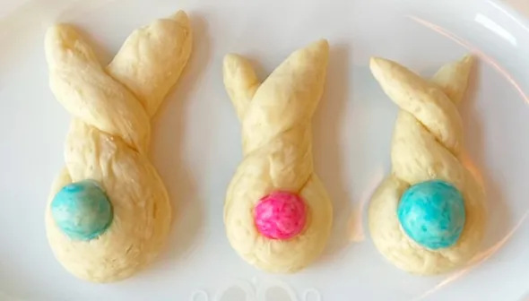 Three pieces of specialty bread sit on a white, oval plate. The dough of the bread has been twisted into the approximate shapes a rabbit.  Each rabbit has coloured dough ball to represent the tail.