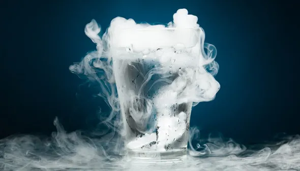A glass sits on a table in front of a blue background. The glass is filled with water and there are two solid pieces of ice near the bottom of the glass. Vapour is flowing from the top of the glass.