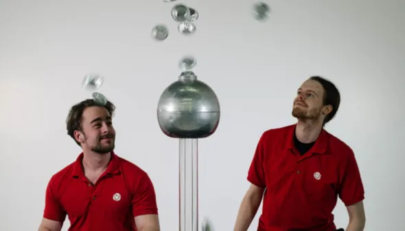 Two men in museum uniforms are on either side of a Van der Graff machine. Metallic disks float in the air, repelled by the machine.
