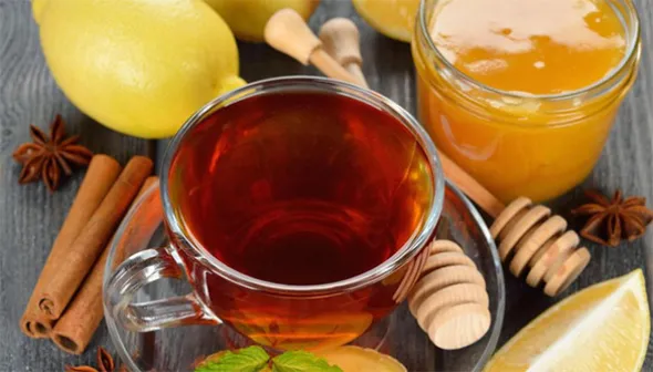 A clear, glass mug filled with tea sits on a matching saucer. It is surrounded by cinnamon sticks, a whole lemon, a lemon wedge, a glass jar filled with honey, two wooden honey dippers, star anise, and two slices of fresh ginger root.
