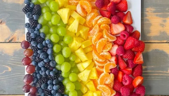An aerial view of a white rectangular platter, covered in a rainbow-coloured assortment of fresh fruit. A small dish at the top holds a white yogurt dip, and a second dish at the bottom holds multiple oatmeal bites.