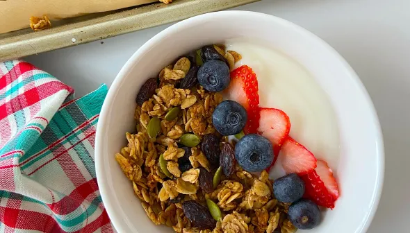 A bowl of granola, yogurt, and fresh fruit sits on a white surface. Next to it, a baking pan and a wooden spoon are filled with the freshly-baked granola.