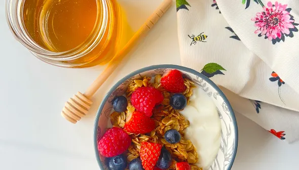 A bowl of granola with mixed berries site next to an open jar of honey, a wooden honey spoon and a floral napkin.
