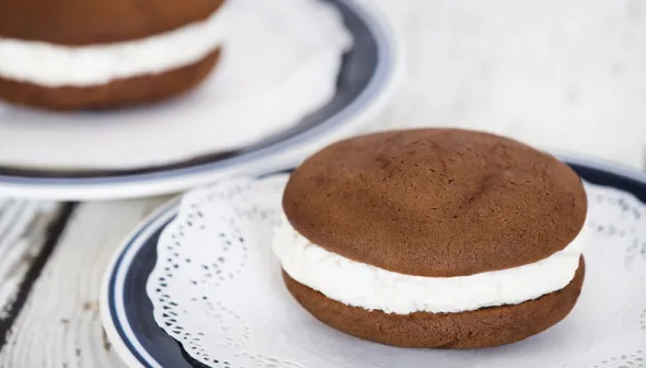 A chocolate cookie sandwich, filled with vanilla ice cream, sits on a small plate on top of a lace doily. A second dessert sandwich is visible in the background.